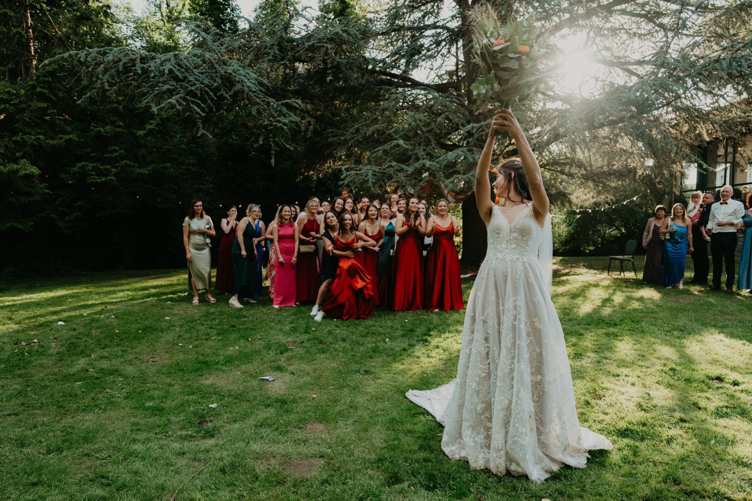 La mariée s’apprête à lancer son bouquet vers ses amies, dans un parc ensoleillé.
