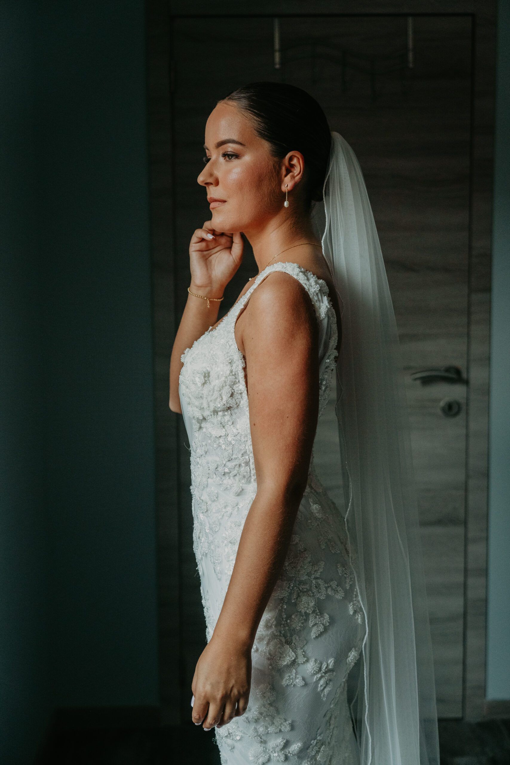 La mariée, en robe de dentelle et long voile, ajuste sa boucle d’oreille, de profil.