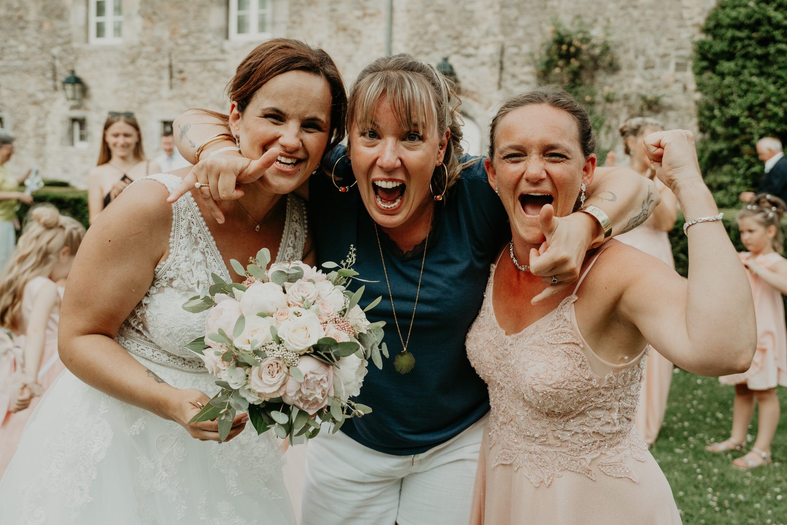 La mariée et ses amies éclatent de rire, bouquet en main, dans le jardin d’un château.